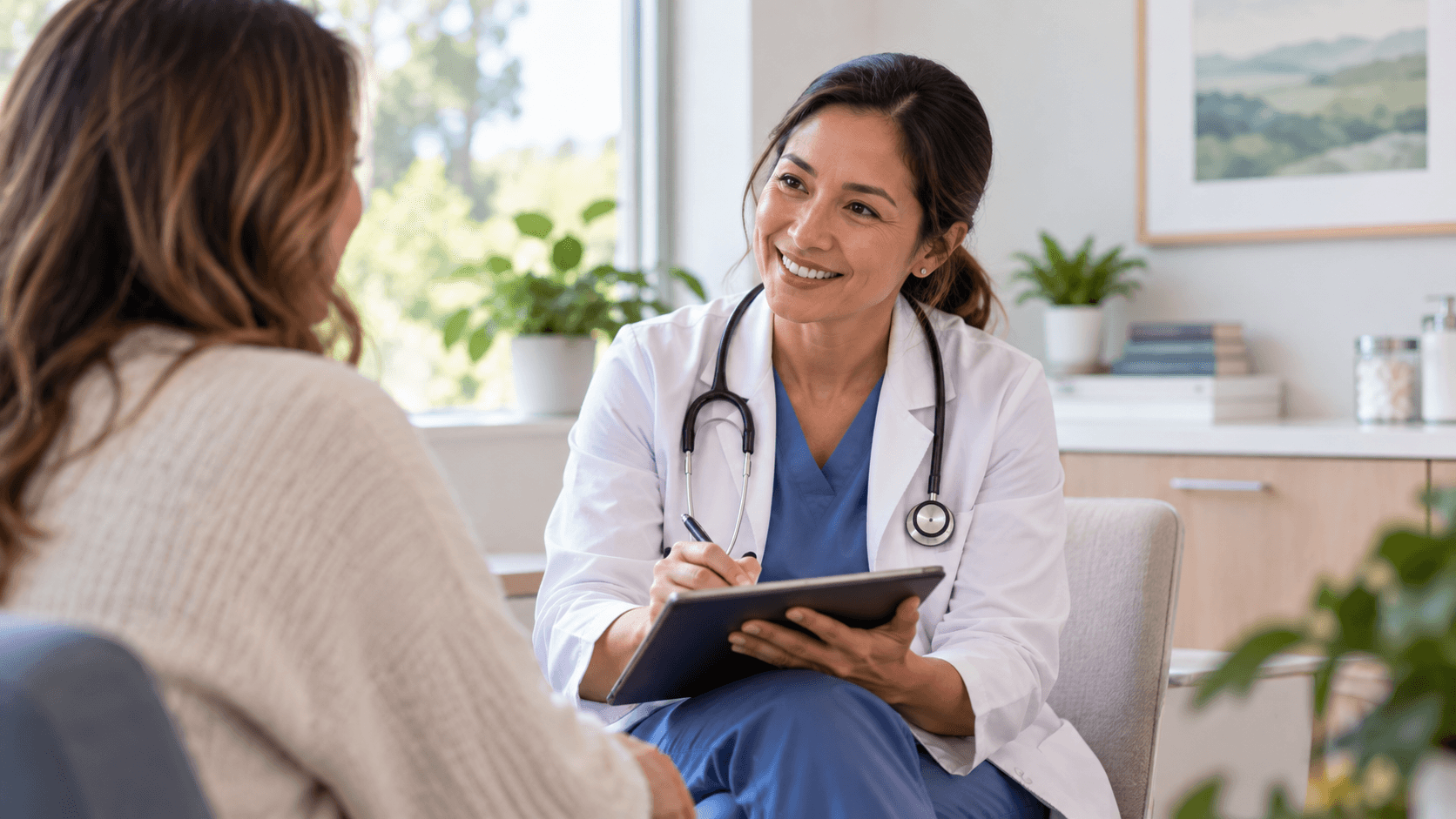 A healthcare provider in scrubs and a stethoscope reviewing notes with a patient in a bright office.